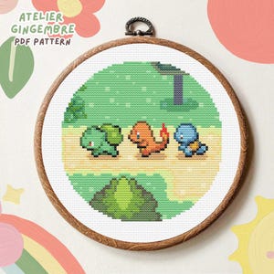 Peut inclure: Modèle de point de croix représentant trois personnages Pokémon : Bulbizarre, Salamèche et Carapuce, marchant sur un chemin dans un décor de forêt verte. Le motif est dans un cercle de broderie en bois rond.