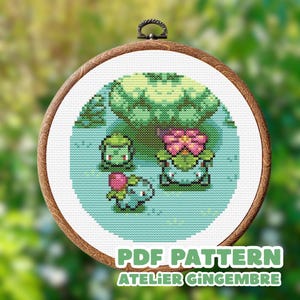 May include: A cross-stitch embroidery featuring three pixelated Pokémon characters in a circular design. The characters are green, pink, and blue, set against a light blue background. The text "PDF PATTERN ATELIER GINGEMBRE" is at the bottom. The embroidery is in a wooden hoop.