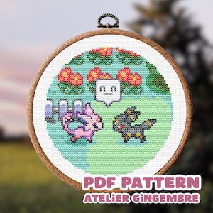 Pode incluir: Um bastidor de bordado circular de madeira contendo um design em ponto cruz de duas pequenas criaturas, uma rosa e uma cinza-escura, num campo verde com flores cor de laranja, uma cerca branca e um balão com o texto PDF PATTERN ATELIER GINGEMBRE.