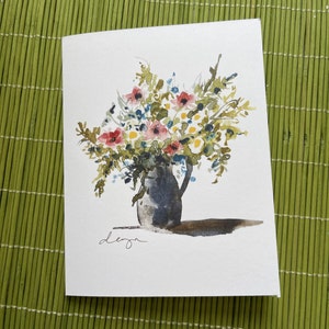 Può includere: Un dipinto ad acquerello di un mazzo di fiori rosa, rossi e bianchi in un vaso di ceramica grigio. I fiori sono disposti in uno stile sciolto e naturale, e il vaso ha un design semplice ed elegante. Il dipinto è firmato "deza" nell'angolo in basso a sinistra.