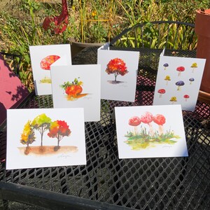May include: Five watercolor greeting cards featuring autumnal themes. The cards depict a pumpkin, a tree with red leaves, a tree with yellow and green leaves, three red mushrooms, and a pattern of red and purple mushrooms.
