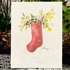 May include: A red Christmas stocking with a white top filled with green and yellow foliage. The stocking is on a white background with the word "design" written in black ink.