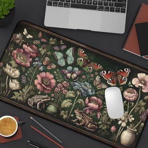 Pode incluir: Tapete de rato retangular com um design botânico com borboletas, flores e rãs em tons de rosa, azul e verde. O tapete tem uma borda marrom escura e está sobre uma superfície escura.