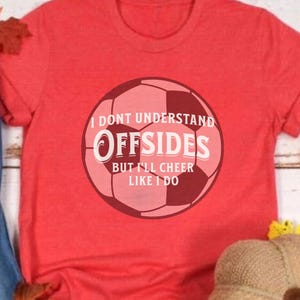 May include: Red t-shirt with a soccer ball graphic. The graphic includes the text "I DON'T UNDERSTAND OFFSIDES BUT I'LL CHEER LIKE I DO". The shirt is displayed with fall-themed decorations, including leaves and a decorative pumpkin.