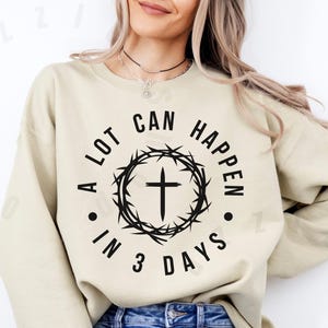 Könnte beinhalten: Ein beiger Pullover mit einem schwarzen Grafikdruck, der "A lot can happen in 3 days." lautet. Die Grafik zeigt ein Kreuz in einem Kreis aus Dornen.