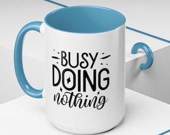 Peut inclure: Une tasse en céramique blanche avec un bord et une anse bleus. La tasse porte une inscription noire qui dit "Busy Doing Nothing".