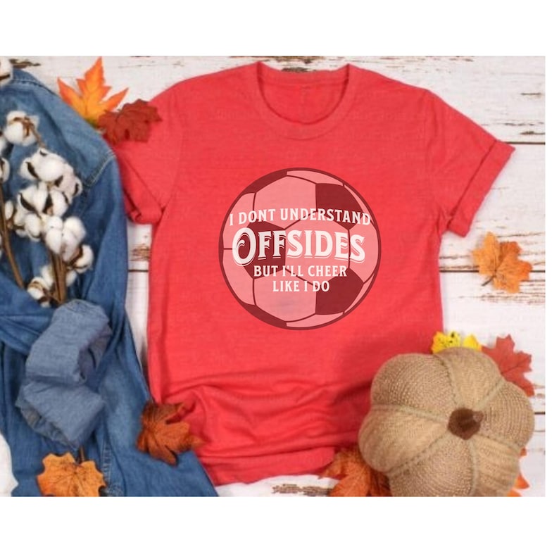 May include: Red t-shirt with a soccer ball graphic. The graphic includes the text "I DON'T UNDERSTAND OFFSIDES BUT I'LL CHEER LIKE I DO". The shirt is displayed with fall-themed decorations, including leaves and a decorative pumpkin.