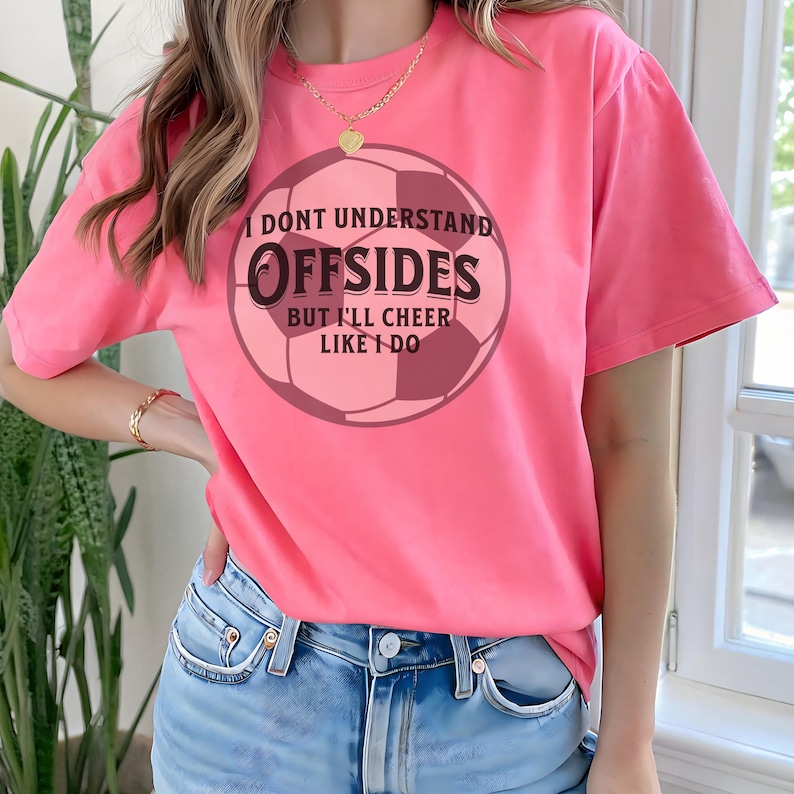 May include: Pink t-shirt with a soccer ball graphic. The graphic includes the text "I DON'T UNDERSTAND OFFSIDES BUT I'LL CHEER LIKE I DO" in a circular design. The shirt is a solid pink color.