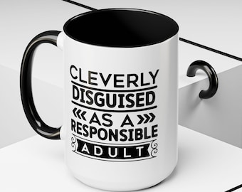 Peut inclure: Une tasse à café en céramique blanche avec un bord et une poignée noirs. La tasse porte une inscription noire qui dit "CLEVERLY DISGUISED << AS A >> RESPONSIBLE ADULT" avec des fioritures décoratives.
