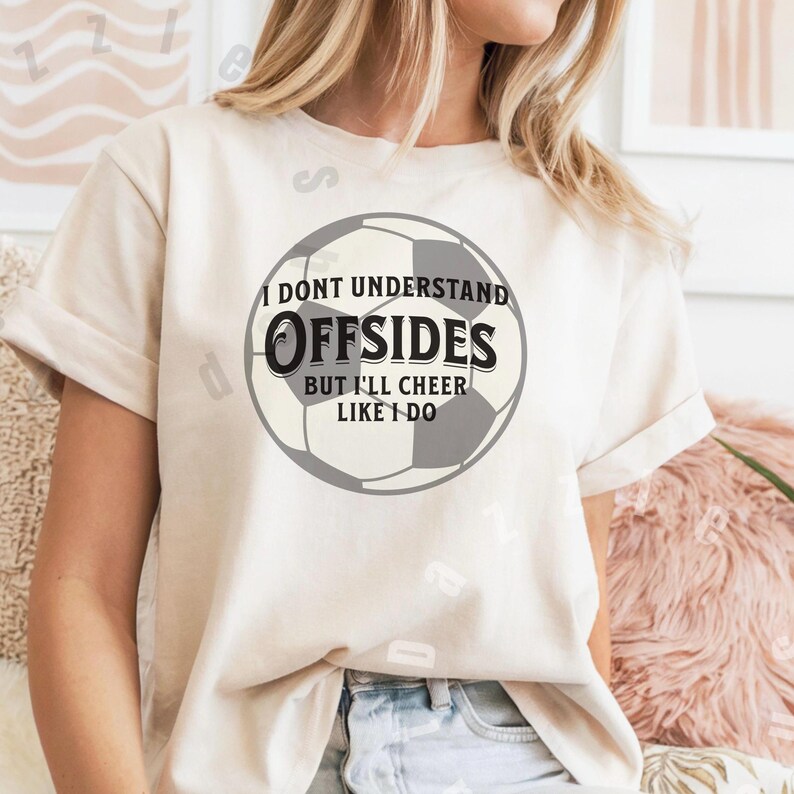 May include: A white t-shirt with a black and gray soccer ball graphic. The text on the shirt reads "I don't understand Offsides but I'll cheer like I do."