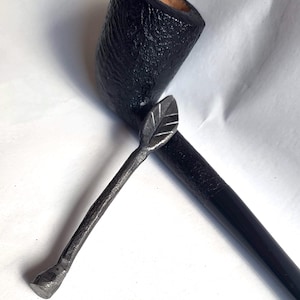 Könnte beinhalten: Eine schwarze Holzpfeife mit einem schwarzen Metallstopfer. Der Stopfer hat ein Blattdesign.