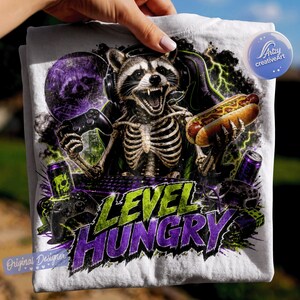 May include: A white t-shirt featuring a graphic of a skeleton raccoon wearing headphones, holding a game controller and a hot dog. The design includes green lightning, a purple circular element, and the text LEVEL HUNGRY in bold purple letters.