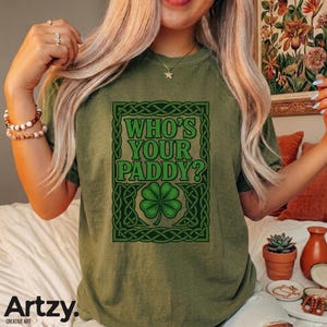May include: Olive green t-shirt with a Celtic knot design and the text "WHO'S YOUR PADDY?" in green. The design includes a four-leaf clover. The shirt is a casual style, suitable for St. Patrick's Day celebrations.