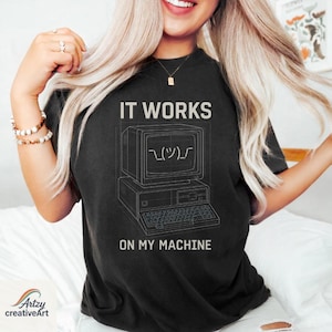May include: Black t-shirt featuring the text "IT WORKS" above a retro computer graphic and the phrase "ON MY MACHINE" below. The computer graphic includes a smiley face. A casual wear item.