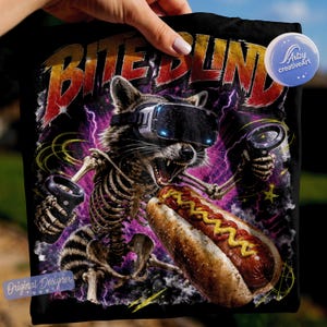 May include: A black t-shirt featuring a graphic of a skeletal raccoon wearing a virtual reality headset and holding controllers. The raccoon is eating a hot dog with mustard. The text BITE BLIND is printed in red and orange above the raccoon.