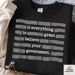 Può includere: Felpa nera con la scritta "everything is great believe your government" in bianco. Il testo è disposto a forma di mattone con rettangoli grigi. La felpa ha uno scollo tondo.