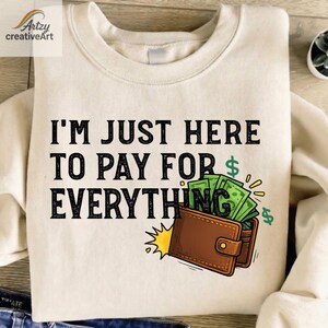 May include: Cream-colored sweatshirt with the text "I'M JUST HERE TO PAY FOR EVERYTHING" in black. A cartoon illustration of a brown wallet overflowing with green dollar bills is below the text.