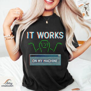 Può includere: T-shirt nera con la scritta "IT WORKS" in un effetto glitch, sopra una grafica verde e una finestra del computer che dice "ON MY MACHINE". La maglietta è di colore scuro e ha un girocollo.