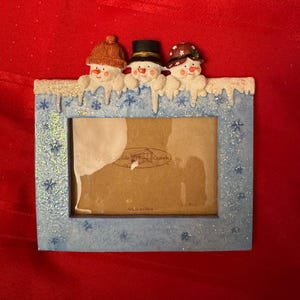 Op de afbeelding: Een rechthoekige fotolijst met een lichtblauwe rand versierd met sneeuwvlokken en glitter. Bovenop staan drie sneeuwpopfiguren met hoeden en wortelneuzen. De lijst heeft een bruine achterkant.