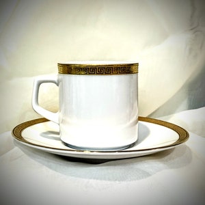 Op de afbeelding: Een wit porseleinen koffiekopje en schotel met een gouden Grieks sleutelmotief rond de rand.
