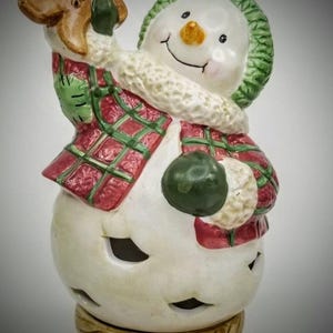 May include: A ceramic snowman figurine with a green knit cap and a red plaid scarf. The snowman holds a gingerbread man and a green ball. The base has a wood-like texture, and the snowman has cut-out eyes and mouth.