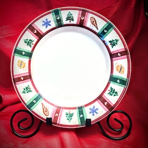 May include: A white ceramic plate with a decorative border featuring Christmas tree, snowflake, and ornament designs in green, red, and blue. The plate is displayed on a black metal stand against a red background.