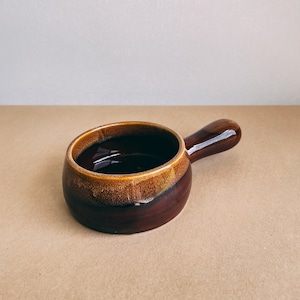 Può includere: Un ramekin in ceramica marrone con un manico. Il ramekin ha una glassa marrone con un bordo marrone chiaro.