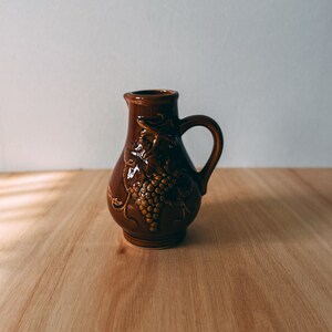 以下が含まれることがあります： ブドウの蔓のデザインが施された茶色の陶器のピッチャー。ピッチャーは持ち手と丸い本体を持っています。