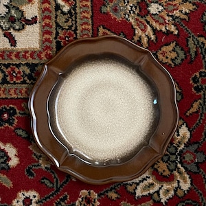 May include: A brown ceramic plate with a scalloped edge. The plate has a speckled, off-white center and a darker brown rim. The plate is likely vintage and suitable for serving food.