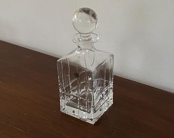 May include: A clear, square-shaped glass decanter with a round stopper. The decanter has vertical and horizontal etched lines, creating a grid pattern. The stopper is a clear, spherical shape. The decanter sits on a dark brown wooden surface.