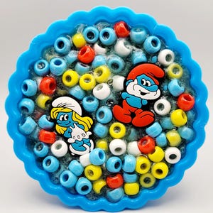 May include: A blue resin coaster with a wavy edge, filled with colorful beads and featuring a Smurf and Smurfette figurine.