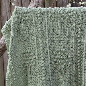 May include: A sage green crocheted blanket with a textured pattern. The blanket is hanging on a wooden fence with a tree branch in the background.