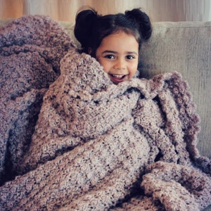 May include: A young girl sits on a couch wrapped in a soft, purple crocheted blanket. The blanket has a textured, chunky stitch pattern. The text "HOLLAND DESIGNS CROCHET" is visible at the bottom of the image.