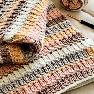 May include: A crocheted blanket featuring horizontal stripes in shades of cream, soft pink, light brown, and grey. The texture consists of repeating raised vertical stitches. A ball of matching yarn and a metal crochet hook are positioned nearby.