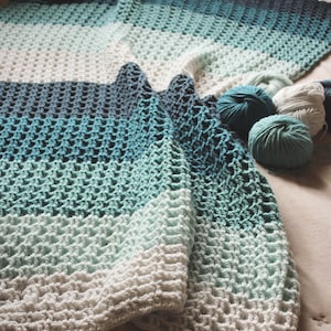 May include: A crocheted blanket with a white and blue ombre pattern. The blanket is made with a textured stitch and is folded on a beige surface. There are three balls of yarn in the colours blue and white.