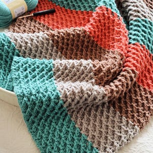 Puede incluir: Una manta de ganchillo texturizada hecha a mano, con un diseño de patchwork. La manta presenta secciones de color turquesa, naranja, marrón y gris. Se ven un ganchillo y lana, lo que sugiere un proyecto artesanal. La manta está doblada, mostrando el detallado trabajo de puntadas.