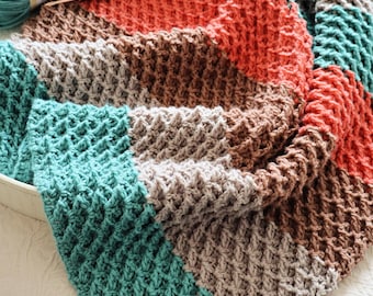 May include: A handmade, textured crochet blanket in a patchwork design. The blanket features sections of teal, orange, brown, and gray. A crochet hook and yarn are visible, suggesting a craft project. The blanket is folded, showcasing the detailed stitch work.