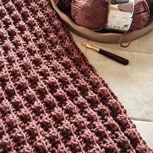 May include: A dusty rose crochet blanket featuring a textured waffle stitch pattern, accompanied by several matching yarn balls labeled hue + me by Two of Wands and a dark wooden crochet hook with a gold-colored metal tip resting nearby.
