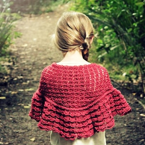 May include: A young person wearing a red crocheted shawl with a scalloped edge. The shawl is worn over a white shirt and blue jeans.