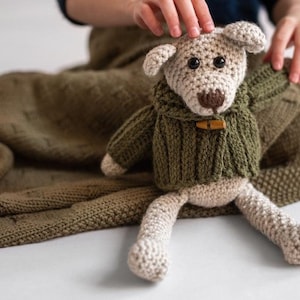 Puede incluir: Un perro de peluche tejido a crochet que lleva un suéter verde con un botón de madera. El perro está sentado en una manta de punto verde.
