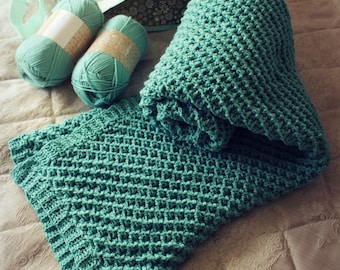 May include: A teal crocheted blanket with a textured waffle stitch pattern. Two skeins of teal yarn are in the background.