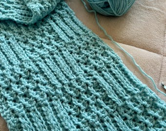 Op de afbeelding: Een licht turquoise gehaakt stoffen stuk met een gestructureerd ribbelpatroon met verticale kolommen van steken. Een bijpassende bol licht turquoise garen bevindt zich in de rechterbovenhoek, met een enkele draad die naar beneden loopt.