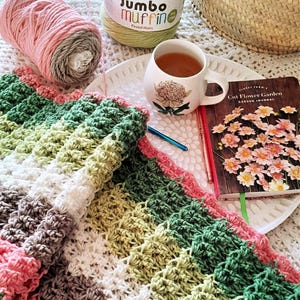 May include: A crocheted blanket with stripes of pink, green, white, and brown yarn. The blanket is folded on a white surface with a cup of tea and a book titled "Cut Flower Garden" on a white plate.