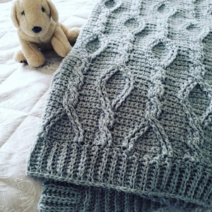 May include: A gray crocheted blanket with a cable knit pattern. The blanket is folded in half and has a soft, textured surface.