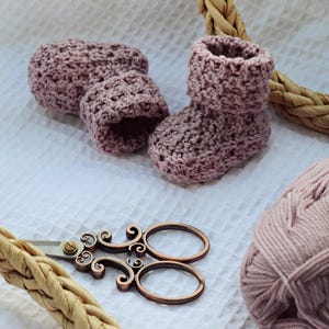 Könnte beinhalten: Ein Paar gehäkelte Babyschuhe in einem staubigen Rosaton, mit einem Knäuel Garn und einer Schere mit Verzierungen auf weißem Hintergrund.