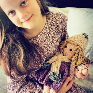 May include: A handmade crochet doll with a beige hat, blonde yarn hair, and a purple dress. The doll has a beige scarf and is held by a person wearing a floral dress.