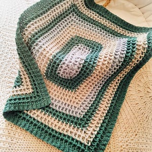 Könnte beinhalten: Eine handgefertigte Häkeldecke in Grün-, Beige- und Lavendeltönen. Die quadratische Decke hat ein strukturiertes Muster mit einem zentralen quadratischen Design und einem durchgehenden grünen Rand. Die Decke ist über eine weiße Oberfläche gefaltet.