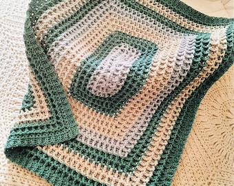 May include: A handmade crochet blanket in shades of green, beige, and lavender. The square blanket features a textured pattern with a central square design and a solid green border. The blanket is folded over a white surface.