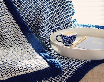 May include: A blue and white knitted throw blanket with a dark blue border, draped over a chair with a blue and white patterned pillow. A white tray with a blue and gold patterned mug and a notebook is on the chair.