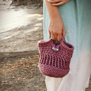 May include: A close-up of a crocheted tote bag in a dusty rose color. The bag has a simple design and a light blue plastic clasp.